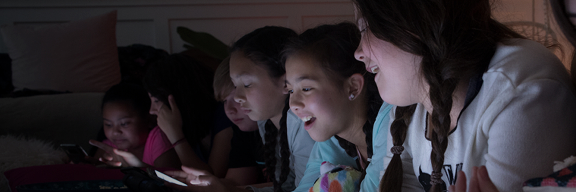 A group of pre-teens girls areat a slumber party. They are all on their devices, laughing and sharing images.