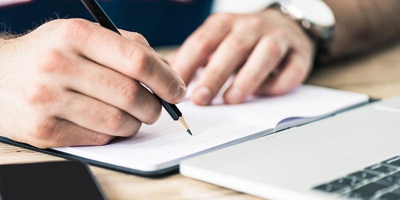 Person's hands writing in notebook in front of laptop