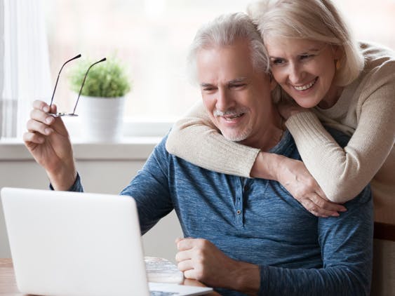 An older Caucasian couples laugh as they FaceTime on a laptop.