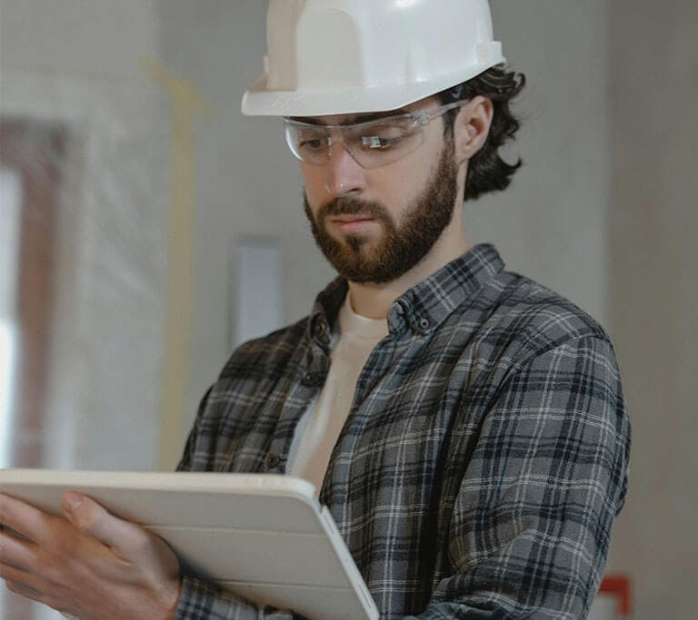 A person wearing a white hard hat and a grey, checkered button-up shirt is standing in a room that’s being renovated while holding a tablet.