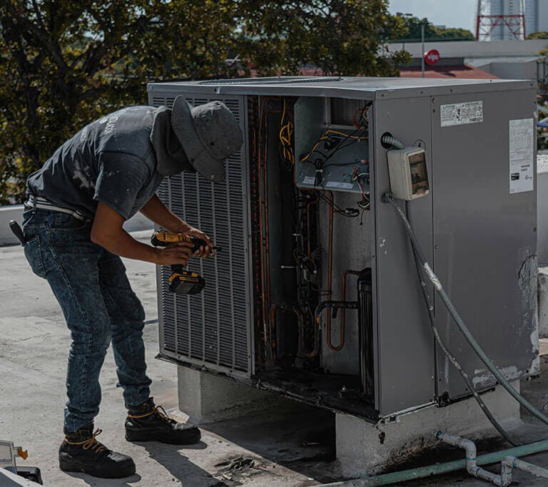 An HVAC professional wearing a grey shirt, jeans, work boots and a grey bucket hat holds a black and yellow drill up to a very large, silver HVAC unit.