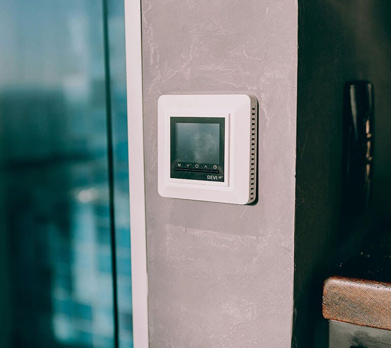 A white smart thermostat on a grey wall next to a gate and a window.