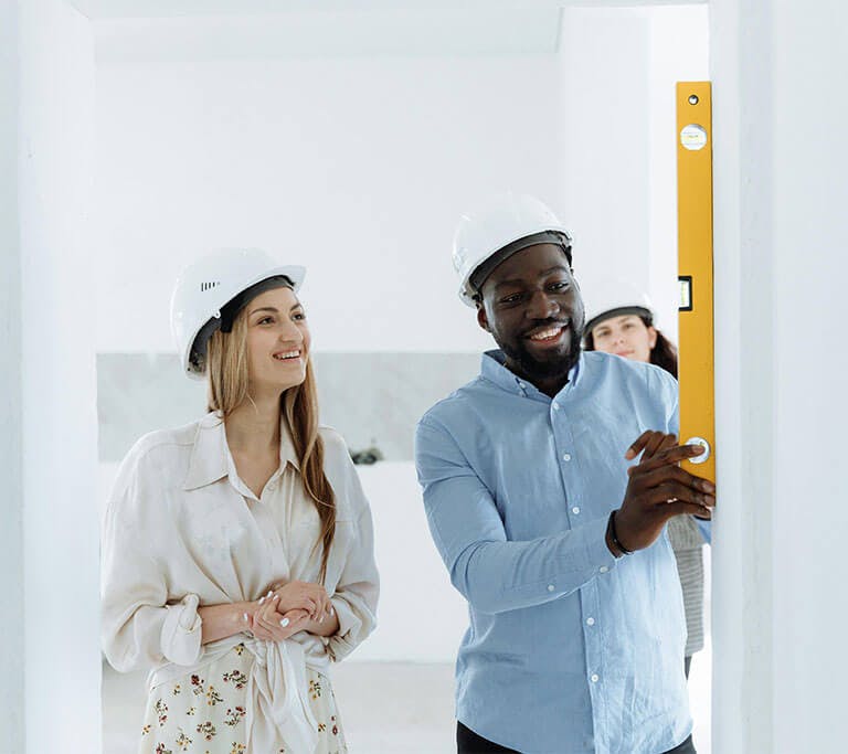Three people wearing hard hats review a wall measurement using a level tool inside a bright, unfinished space.