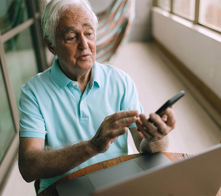 An older man sits on his screened-in porch using a mobile phone and a laptop.