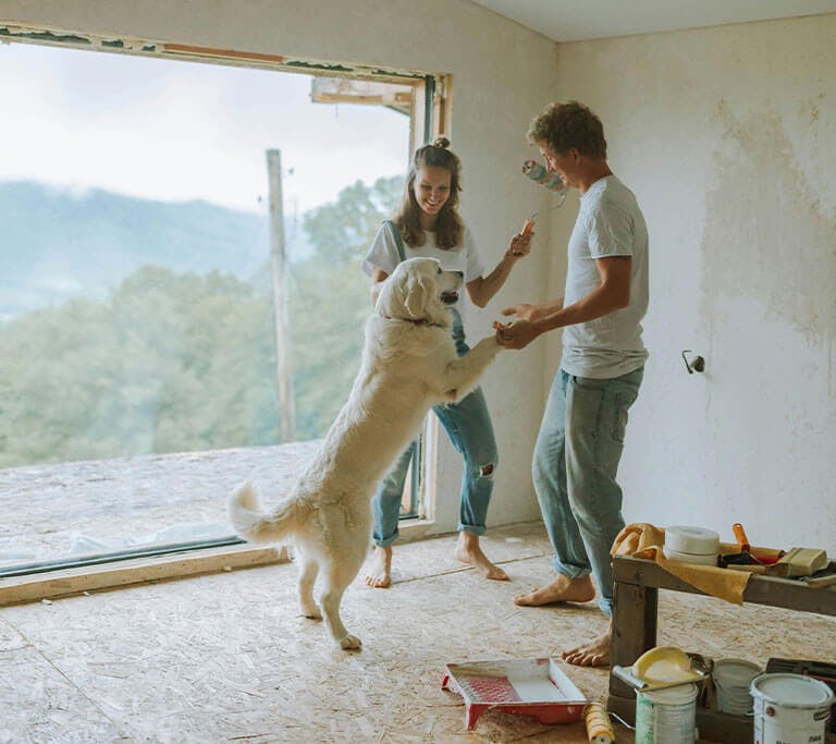 Two people wearing white shirts and jeans take a break from renovating to dance happily with their golden retriever in a room with unfinished floors and walls.