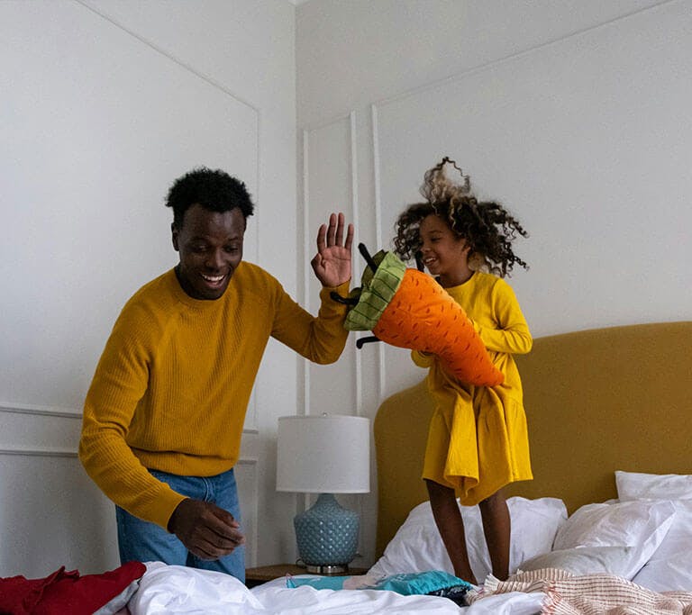 A father laughs while his smiling daughter jumps on a bed while holding a carrot toy in a white room.