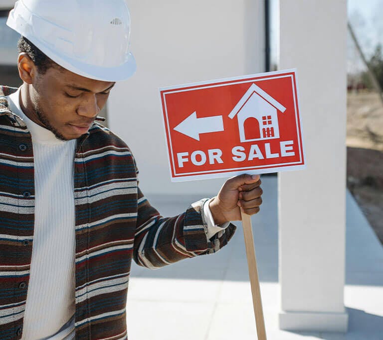 A man wearing a knit sweater and a stripped jacket holds a “For Sale” sign in front of a beautiful white house.