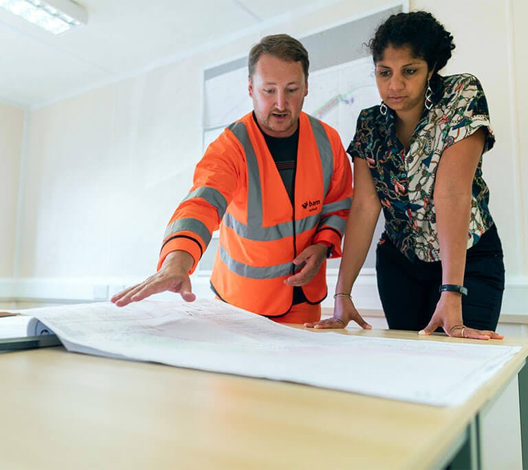 A homeowner talks to a professional wearing a high-visibility safety jacket as they look at a blueprint on a table.