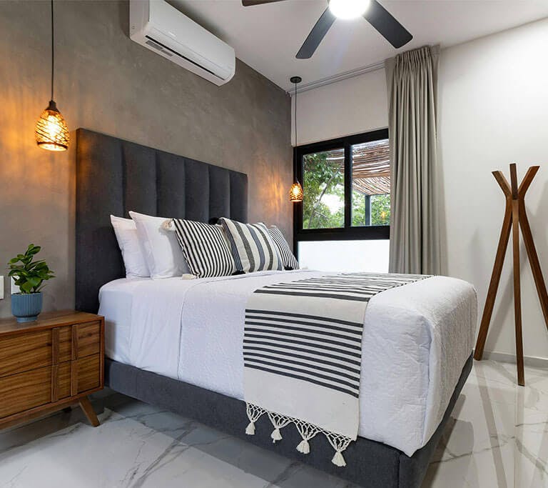 A modern bedroom with a wooden sculpture, a window, grey walls, a marbled floor, wooden side table, houseplant and neatly made bed with a mini split AC unit mounted near the ceiling.