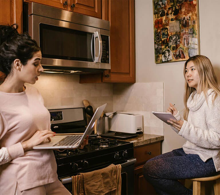 A person holds a notebook and pen while talking to another person holding a laptop in a kitchen with wooden cabinets and white walls near a microwave and thick walls.