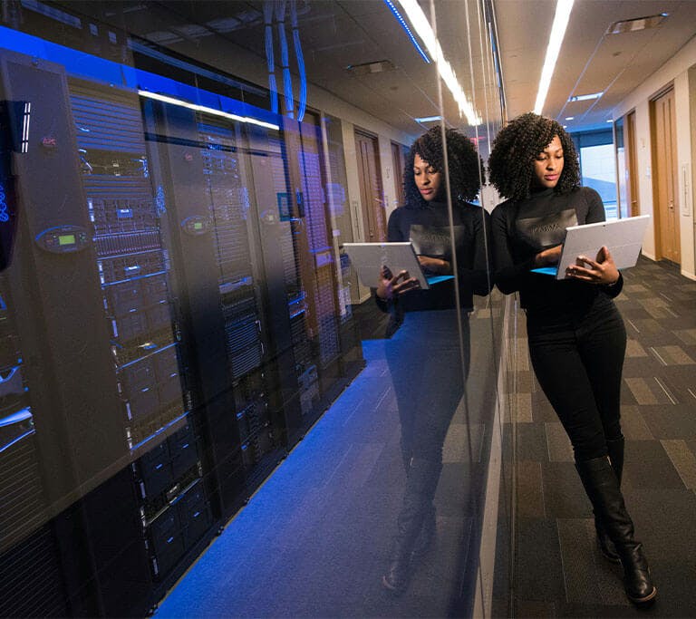 A person wearing a stylish all-black outfit holds a laptop next to a futuristic server room.