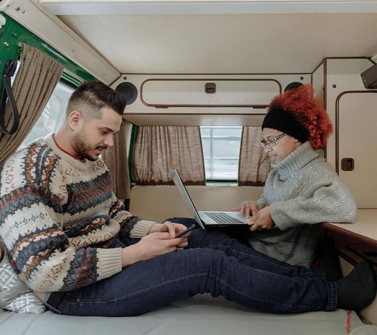 A couple wearing sweaters lounge in a camper van while using their devices.