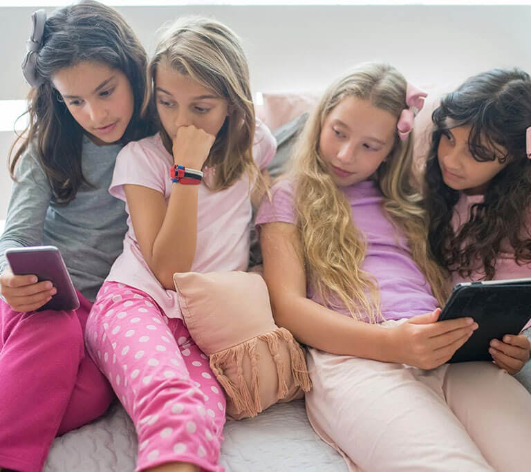 Four children sit together on a bed with pillows, watching content on a phone and a tablet.