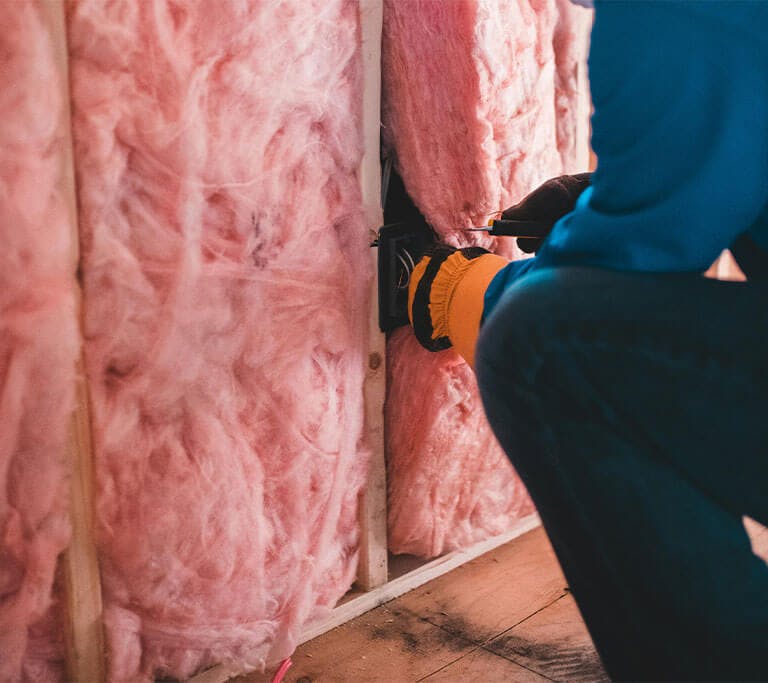 A person wearing jeans and safety gloves crouches near the floor in an unfinished room while installing pink mineral wool insulation.