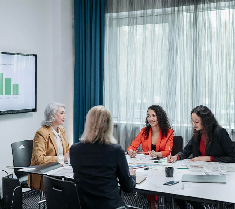 A group meets in a conference room while a bar chart is displayed on a wall-mounted screen.
