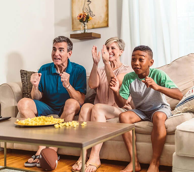 A family of three cheers while sitting on a couch and watching TV in front of a coffee table, which holds two remote controls and a snack plate.