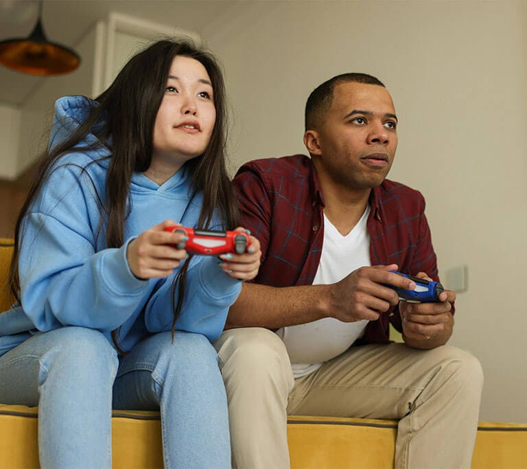 A couple sitting on a yellow couch leans forward, each holding a controller in their hands, as they focus on a video game they’re playing.
