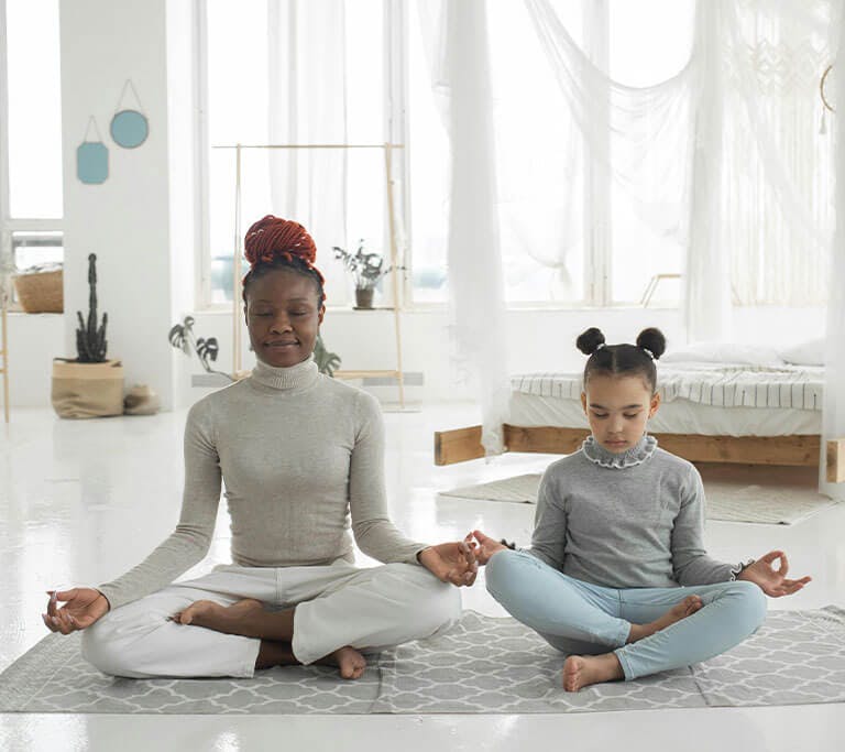A child and adult sit on a mat while meditating, inhaling and exhaling the air inside their home.