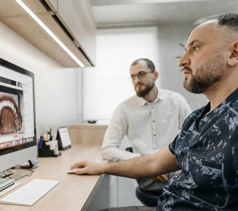 Two people sit at a desk in a dental office, viewing a detailed image of teeth on a large computer screen. One person operates the mouse while the other observes. The environment is clean and modern, with dental tools and notes nearby.