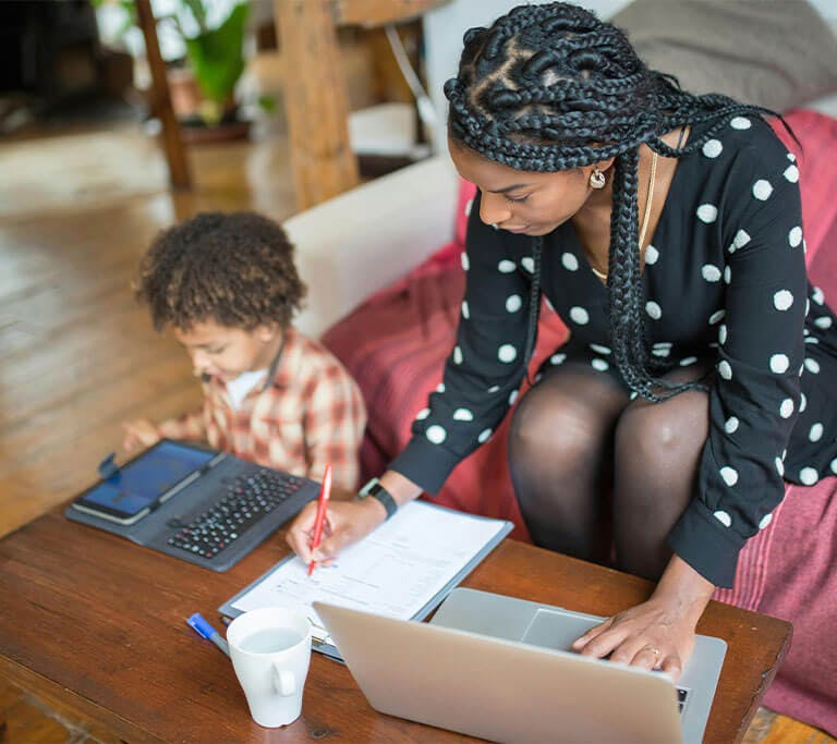 A mother uses a laptop and takes notes while sitting on a couch next to her child who is using a tablet and sitting on the floor.