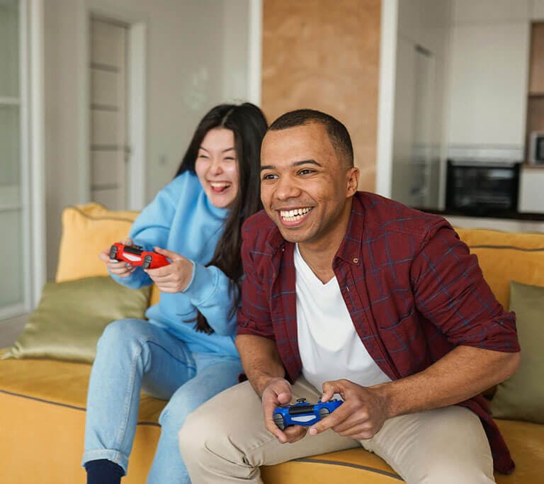 A smiling couple appears to be fully immersed in a video game while sitting on a modern, yellow couch in their apartment, each holding a controller.