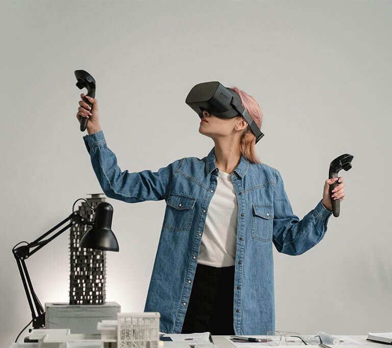 A person with pink hair in a denim jacket and white shirt stands in a white room in front of a work desk while using VR technology.