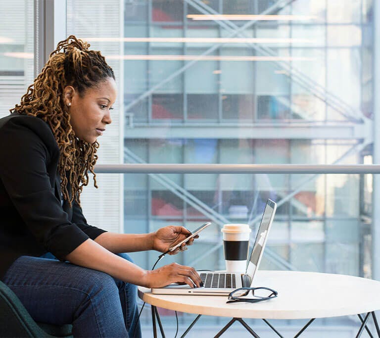 A woman wearing jeans and a black shirt sits at a desk using a laptop and phone by large windows in a city on a sunny day.