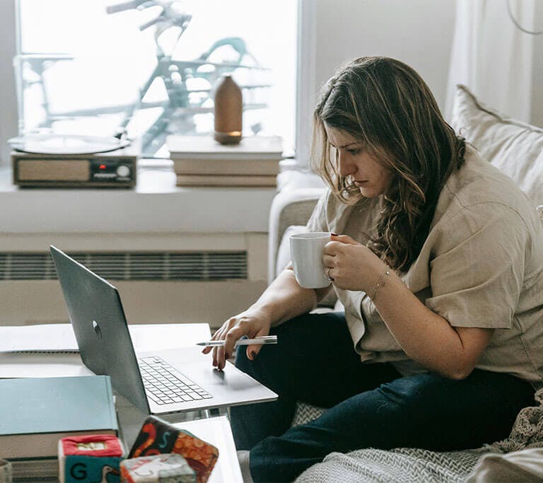 A person holding a pen and mug sits on a couch covered in soft blankets while focusing on using a laptop that’s resting on a coffee table near some books and toys.