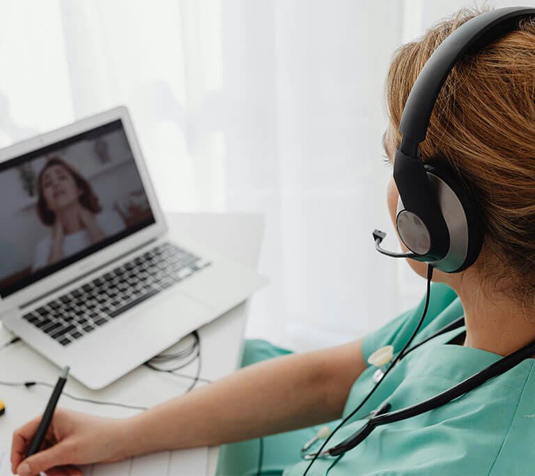 A medical professional wearing green scrubs and a headset takes notes as a person on a laptop screen points to their neck as if they’re describing symptoms.