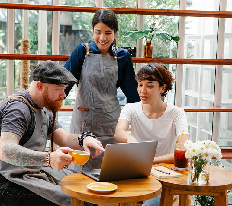 Two café employees in aprons stand and sit around a small round table with a customer, who is seated and using a laptop. One employee holds a yellow coffee cup and gestures toward the screen, while the other smiles and looks on. The café is bright and mod