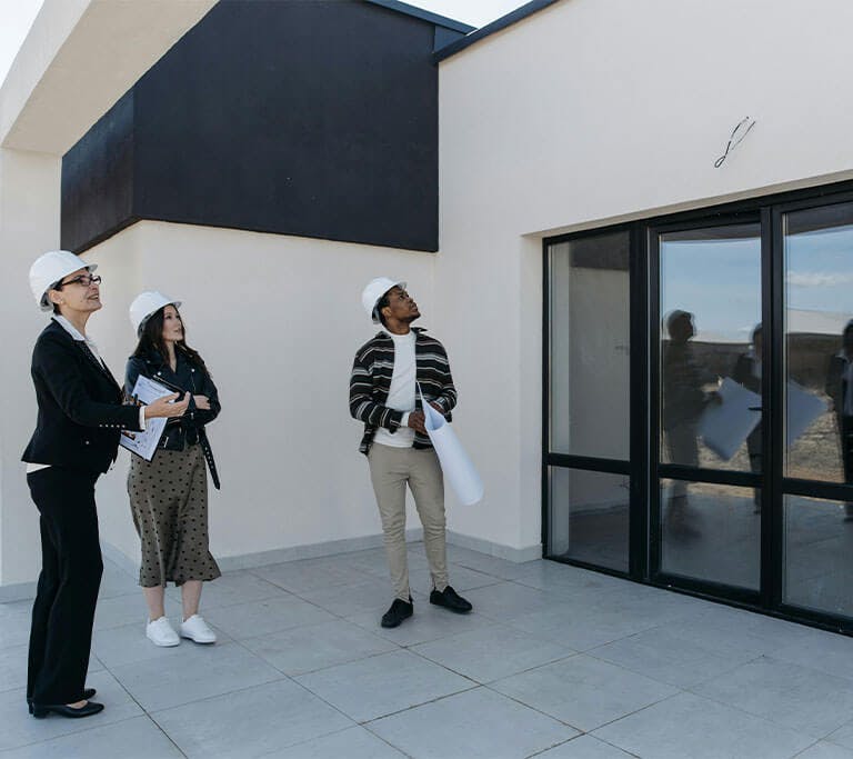 Three people wear white hard hats and stand outside of a modern building, looking at an area of an exterior wall above large windows.