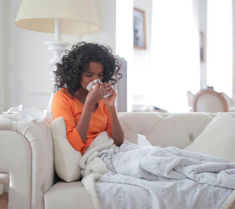 Woman on a couch, under a blanket, sneezes due to poor indoor air quality.