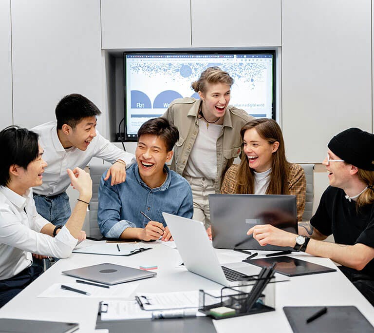 A team of workers smiles and socializes at a meeting room table with various devices, including several laptops and a TV.