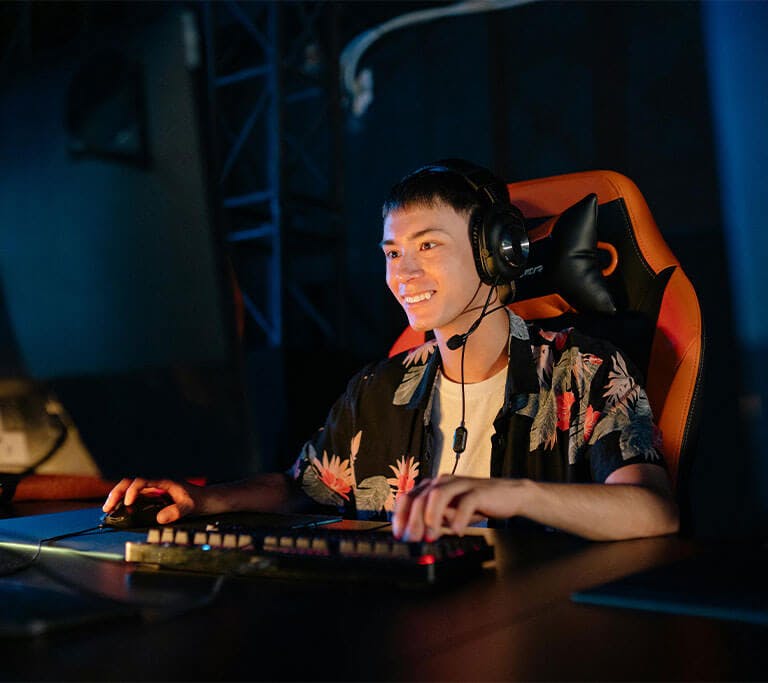 A gamer sits in a red gaming chair at a desk using a keyboard and mouse while smiling at a display.