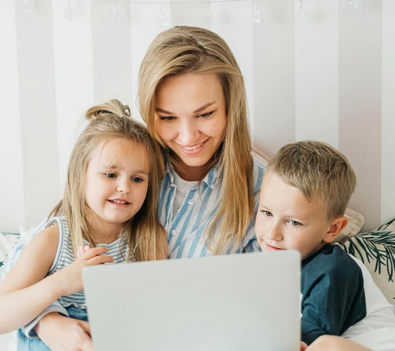 A woman is looking at a laptop with her young son and daughter while smiling.