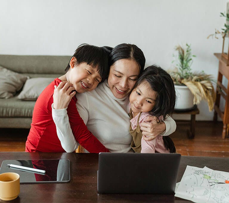 A smiling mother hugs her two children as she sits at a table with an open laptop, tablet, pen stylus and stack of their drawings.