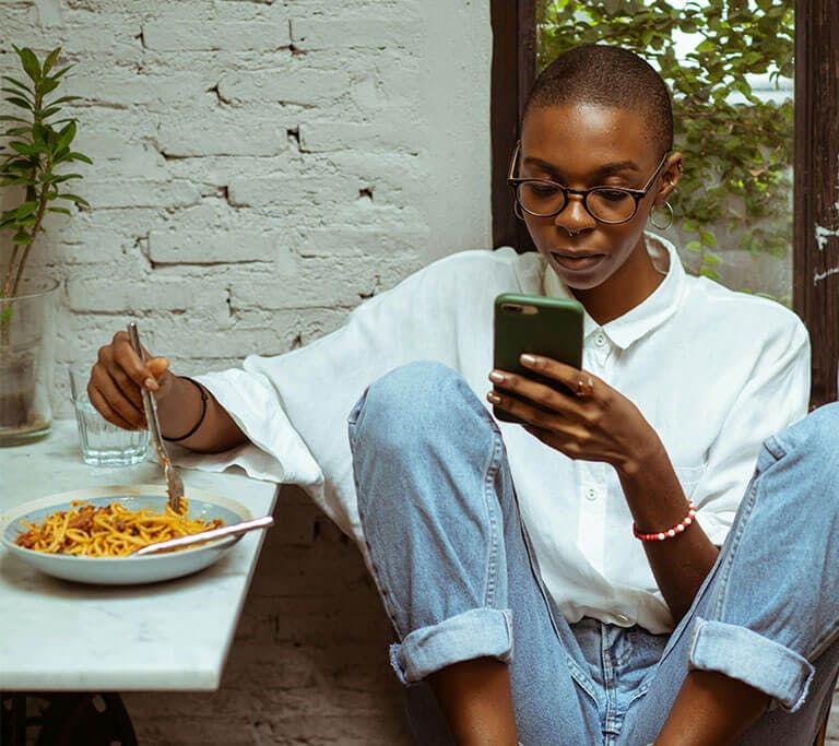 A person wearing glasses eats spaghetti at a table near white brick walls while using their green smart phone.
