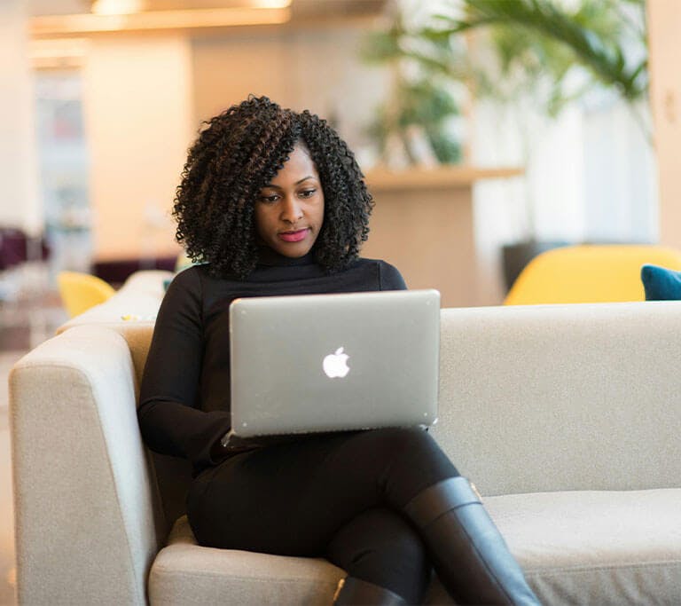 A person wearing black clothes and boots with curly hair sits on a cream couch using her silver Macbook in a white room with a houseplant in the background.