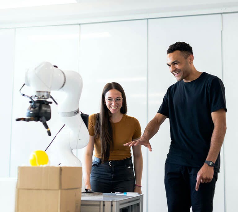 Two engineers smile as they test a white robotic arm they appear to be developing in a white room.
