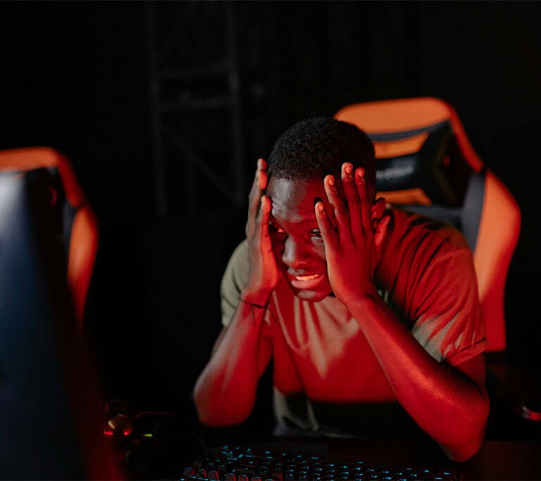 A gamer in a brown t-shirt is sitting in an orange gaming chair at a desk with a backlit mechanical keyboard and computer display, holding their face in their hands with a frustrated or upset expression.