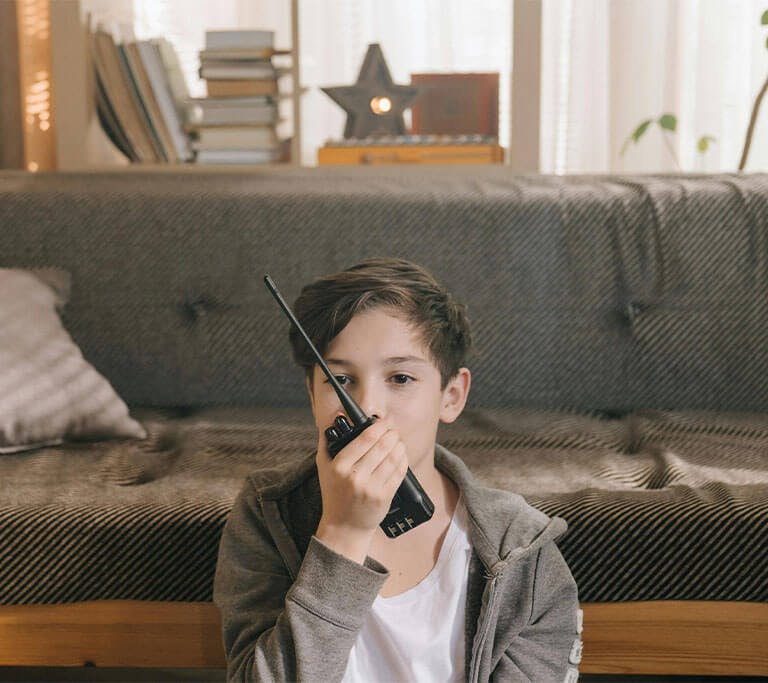A child with short hair and a grey hoodie sits in front of a futon and speaks into a black walkie-talkie.