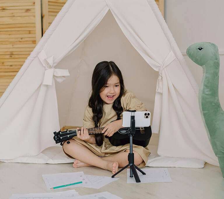 A smiling child wearing a beige dress sits in front of a small, white, toy tent and plays a black ukelele while looking at a white phone on a tripod stand next to a green dinosaur toy.