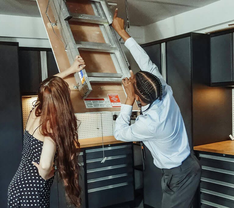Two nicely dressed people pull down an attic ladder from a door in the ceiling of a workshop area.