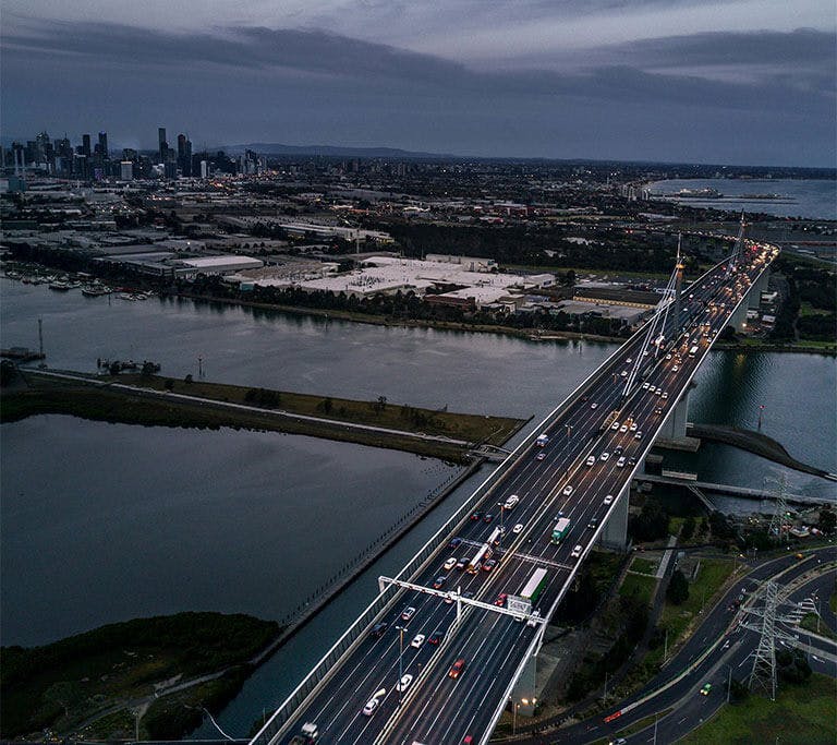 Aerial view of a busy multi-lane bridge crossing water toward a distant city skyline at dusk.