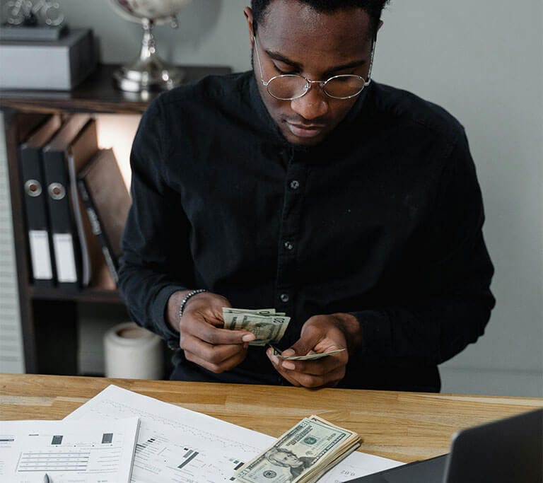 A person in a black shirt wearing round glasses in a beige room counts money while sitting at a desk with a laptop, papers, cash and a calculator.