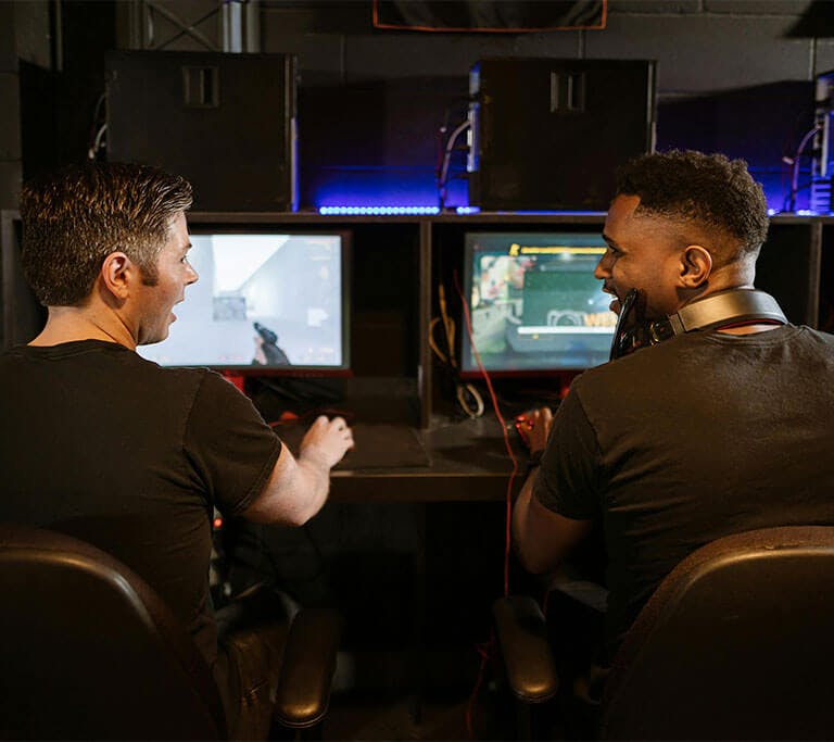 Two gamers smile at each other while playing video games on screens in front of them, with towers in the background glowing blue.