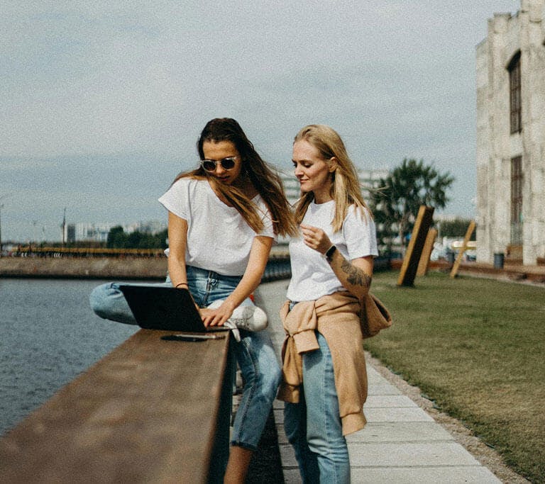 Two adult people in jeans and white shirts sit by the water in a rural area, using a laptop on a cloudy day.