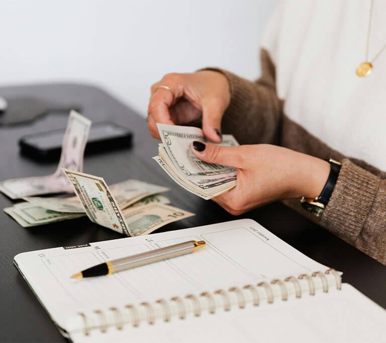 A person counts money with a planner and pen in front of them on a black desk.