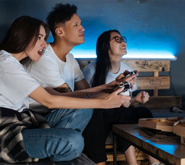 Three friends sit around a wooden table enjoying some pizza as they play video games.