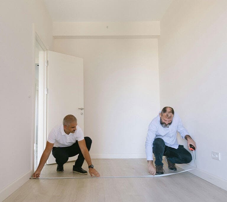 Two people crouch near the ground in a room with white walls and work together to measure the distance between walls with a measuring tape.